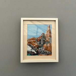 Könnte beinhalten: Aquarellmalerei in einem weißen Rahmen, die eine rustikale Stadt inmitten brauner Felsformationen zeigt. Ein Zug und Gebäude mit roten Dächern sind zu sehen. Der Himmel ist blau.
