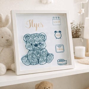 Peut inclure: Un cadre carré blanc contenant une silhouette d'ours bleu remplie de petites roses en papier. Le prénom Ilyes est écrit en lettres dorées. Des icônes indiquent la date 13/12/25, l'heure 2h13, le poids 2148 kg et un ruban à mesurer.