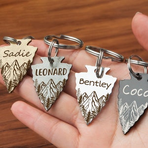 May include: Four silver arrowhead keychains with mountain and tree designs. Each keychain has a different name engraved on it: Sadie, Leonard, Bentley, and Coco.