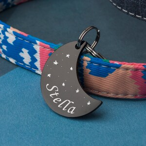 K&ouml;nnte beinhalten: Schwarzer, sichelf&ouml;rmiger Haustier-ID-Anh&auml;nger mit dem Namen "Stella", der in wei&szlig;er Schrift graviert ist. Der Anh&auml;nger ist mit kleinen wei&szlig;en Sternen verziert.