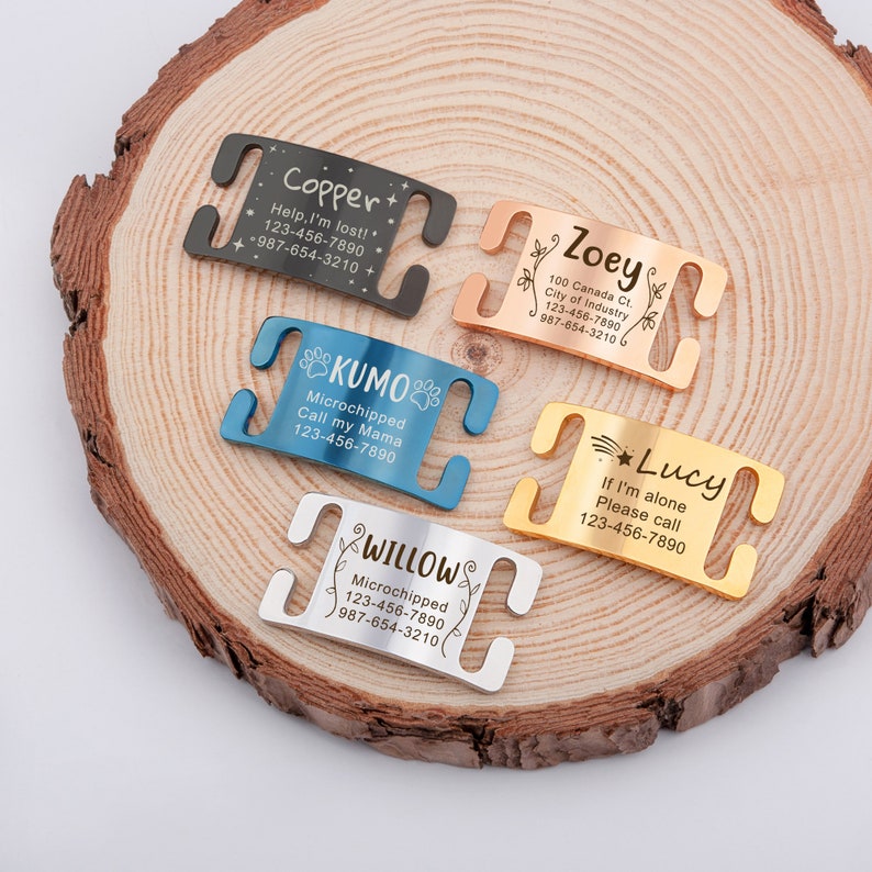 Op de afbeelding: Vijf rechthoekige metalen naamplaatjes voor huisdieren met zijsleuven voor bevestiging aan een halsband. De plaatjes zijn zwart, ros&eacute;goud, blauw, goud en zilverkleurig, elk gegraveerd met een naam, contactgegevens en decoratieve iconen.