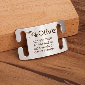 Op de afbeelding: Een zilverkleurig metalen naamplaatje voor huisdieren in een gebogen rechthoekige vorm met vier zijdelingse sleuven. Het plaatje bevat een gegraveerde vallende ster, de naam Olive en contactgegevens: 123-456-7890, 987-654-3210, 100 Canada Ct., City of Industry.