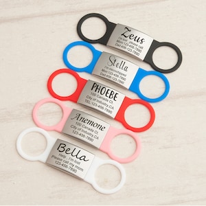 K&ouml;nnte beinhalten: F&uuml;nf bunte Hunde-ID-Tags mit Metallplatten, die mit Namen und Kontaktinformationen graviert sind. Die Tags sind an schwarzen, blauen, roten, rosa und wei&szlig;en Silikon-Schlaufen befestigt.