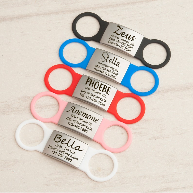 K&ouml;nnte beinhalten: F&uuml;nf bunte Hunde-ID-Tags mit Metallplatten, die mit Namen und Kontaktinformationen graviert sind. Die Tags sind an schwarzen, blauen, roten, rosa und wei&szlig;en Silikon-Schlaufen befestigt.