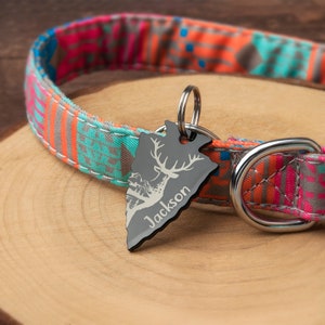K&ouml;nnte beinhalten: Ein schwarzer Metall-Hundeanh&auml;nger in Pfeilspitzenform mit einer Hirsch-Silhouette. Der Anh&auml;nger ist mit dem Namen "Jackson" graviert. Der Anh&auml;nger ist an einem bunten, gemusterten Hundehalsband befestigt.