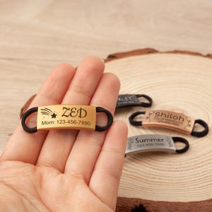 May include: A gold-toned metal identification tag with a black elastic band, engraved with the name ZED, a shooting star graphic, and the text Mom: 123-456-7890. Additional tags in black, rose gold, and silver are visible in the background.