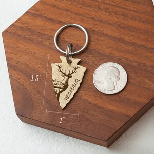 K&ouml;nnte beinhalten: Ein goldfarbener Pfeilspitzen-Schl&uuml;sselanh&auml;nger mit einer Rentier-Silhouette und dem Wort "REINDEER" eingraviert. Der Schl&uuml;sselanh&auml;nger ist 1,5 Zoll hoch und 1 Zoll breit.