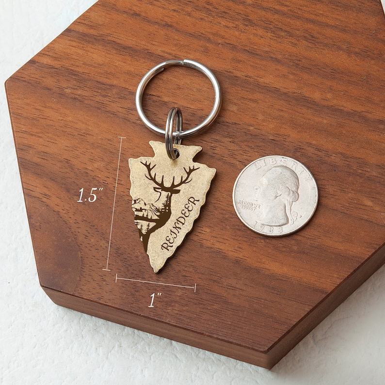 K&ouml;nnte beinhalten: Ein goldfarbener Pfeilspitzen-Schl&uuml;sselanh&auml;nger mit einer Rentier-Silhouette und dem Wort "REINDEER" eingraviert. Der Schl&uuml;sselanh&auml;nger ist 1,5 Zoll hoch und 1 Zoll breit.