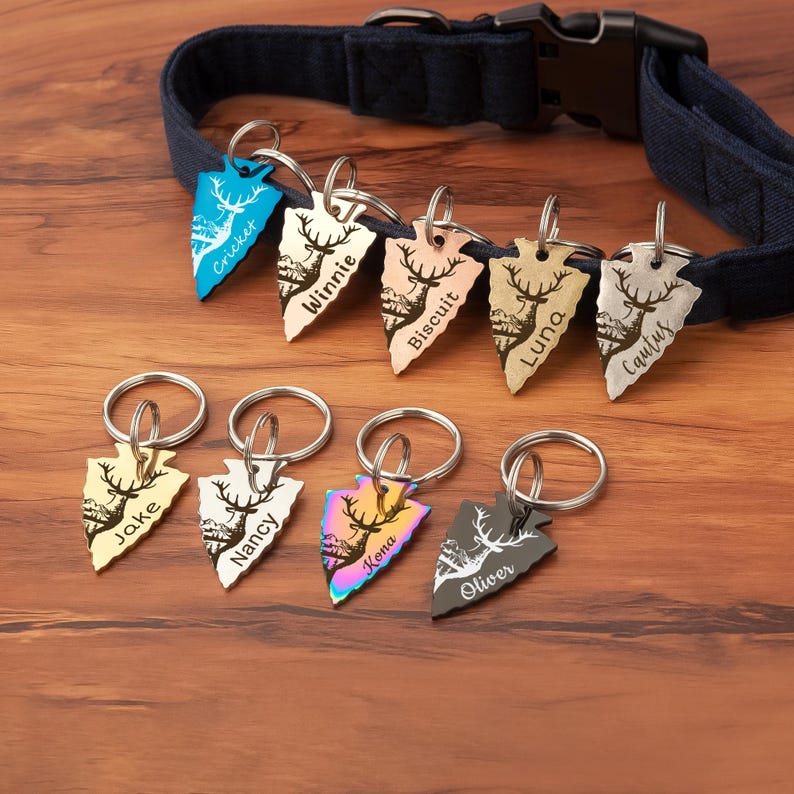 K&ouml;nnte beinhalten: Neun personalisierte, pfeilf&ouml;rmige Hundeanh&auml;nger mit gravierten Namen, darunter Cricket, Winnie, Biscuit, Luna, Cactus, Jake, Nancy, Kona und Oliver. Die Anh&auml;nger sind an Schl&uuml;sselringen befestigt und auf einer Holzoberfl&auml;che angeordnet.