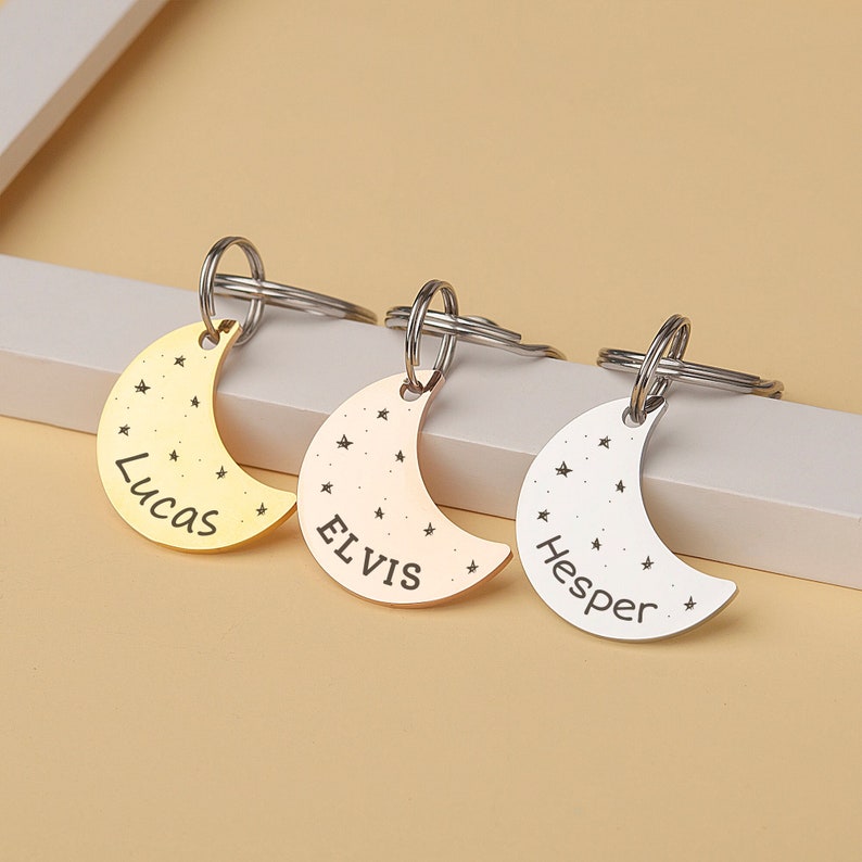 May include: Three silver crescent moon keychains with star accents. The keychains are engraved with the names Lucas, Elvis, and Hesper.
