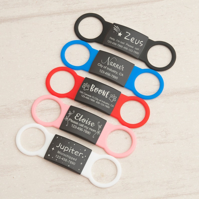 May include: Five different dog ID tags with black metal plates and colorful silicone rings. The tags feature engraved names, phone numbers, and additional information like "Help, I'm lost" and "Microchipped".