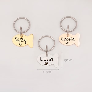 May include: Three gold, silver, and rose gold fish-shaped pet ID tags with key rings. The tags are engraved with the names "Suzy", "Luna", and "Cookie". The tags measure 1 3/16 inches long and 13/16 inches wide.