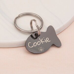 May include: Black metal fish-shaped pet tag with a silver ring and a small bee design. The tag is engraved with the name "Cookie".