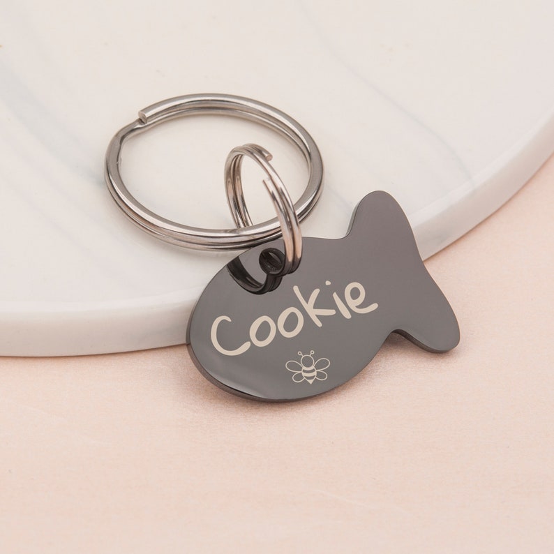May include: Black metal fish-shaped pet tag with a silver ring and a small bee design. The tag is engraved with the name "Cookie".