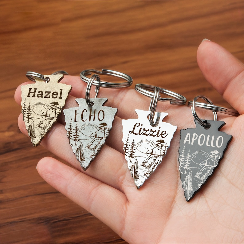 Może przedstawiać: Cztery srebrne breloki w kształcie grotu strzały z wygrawerowanymi imionami i sceną g&oacute;rską. Imiona to Hazel, Echo, Lizzie i Apollo.