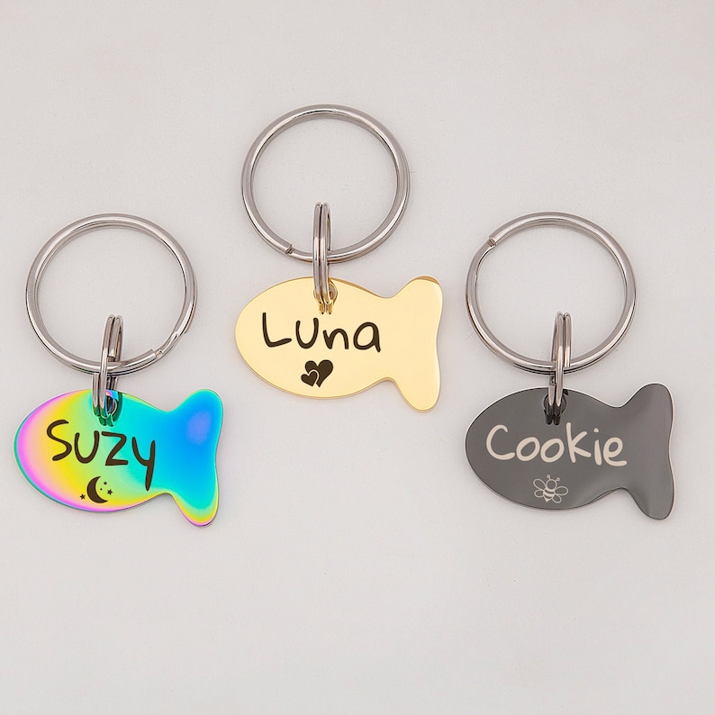 May include: Three gold, silver, and iridescent fish-shaped pet ID tags with key rings. The tags are engraved with the names "Luna", "Suzy", and "Cookie".