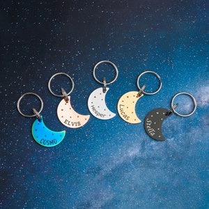 May include: Five silver crescent moon keychains with different colored finishes and engraved names: Cosmo, Elvis, Hesper, Lucas, and Stella.