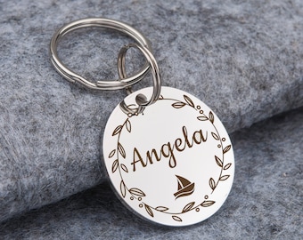 Peut inclure: Porte-clés argenté avec une étiquette ronde ornée d'une couronne de fleurs et le nom "Angela" gravé en cursive. Une petite icône de voilier est centrée dans la couronne.