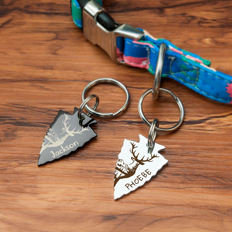K&ouml;nnte beinhalten: Zwei silberne, pfeilspitzenf&ouml;rmige Hundemarken mit gravierten Hirsch-Silhouetten und den Namen "Jackson" und "Phoebe". Die Marken sind an silbernen Schl&uuml;sselringen befestigt.
