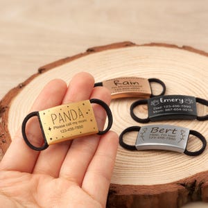 May include: Four rectangular pet ID tags with black elastic bands for collars. One gold tag reads PANDA, Please call my mom, 123-456-7890. One rose gold tag reads Rain, I'm Microchipped, 123-456-4856. One black tag reads Emery, Dad, Mom. One silver tag reads Bert, Help, I'm lost!, 123-456-7890.
