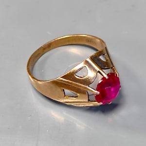 Può includere: Un anello color oro con una grande gemma rotonda di colore rosso brillante. L'anello presenta un design traforato decorativo sui lati. La gemma è incastonata in un'incastonatura a griffe.