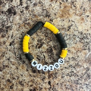 May include: A beaded bracelet featuring alternating segments of flat, circular black and yellow clay beads, accented with small gold-toned metallic spacers. The bracelet includes white spherical beads with black lettering that spell out the word MIZZOU.