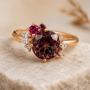 May include: A gold ring featuring a large, round, dark red gemstone as the focal point. Accenting the main stone are smaller, pink and white gemstones. The ring is set on a light-colored, textured surface.