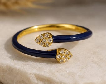 May include: A gold-toned ring with a navy blue enamel band. The ring features two teardrop-shaped accents encrusted with small, clear gemstones. The ring is displayed on a white marble surface.