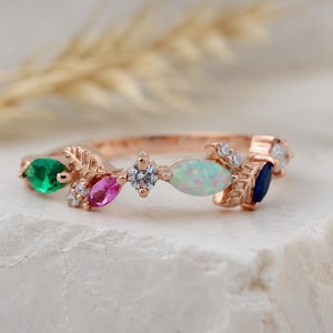 Könnte beinhalten: Ein roségoldener Ring mit Blattdesign, besetzt mit einem grünen Smaragd, einem rosa Rubin, einem weißen Opal, einem blauen Saphir und kleinen Diamanten. Der Ring liegt auf einer weißen Oberfläche. Die Edelsteine werden in Millimetern gemessen.