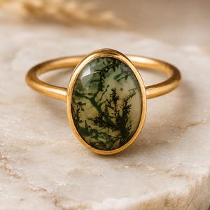 May include: Gold ring featuring an oval moss agate stone. The stone has a translucent appearance with green and white inclusions, set in a gold band. The ring is displayed on a marble surface, showcasing the jewelry's design and color.