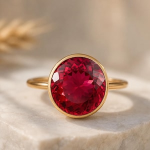 May include: A gold ring featuring a large, oval-cut, deep red gemstone. The gemstone is set in a gold bezel, and the band is a simple, polished gold. The ring is displayed on a white surface.