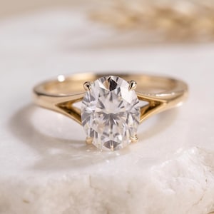 Puede incluir: Un anillo de oro con una gran piedra preciosa transparente de talla ovalada y facetas, montada en un engaste de cuatro garras. La banda presenta un diseño de brazo dividido que se estrecha hacia la piedra central con acabado dorado.
