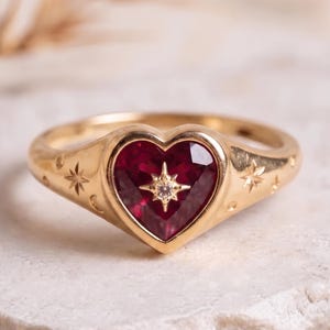 Puede incluir: Un anillo de oro con una piedra preciosa roja en forma de corazón en el centro. La piedra está acentuada con una pequeña incrustación de diamante en forma de estrella. La banda de oro tiene grabados motivos de estrellas y lunas.