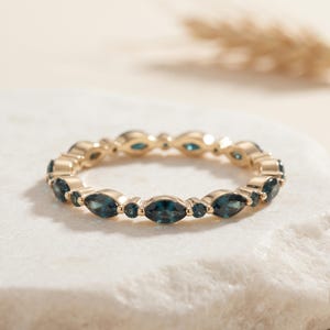 Puede incluir: Un anillo de banda dorada con un patrón alterno de piedras preciosas de color azul oscuro en talla marquesa y redonda. Las piedras están engastadas en una fila continua alrededor de la circunferencia de la banda metálica, creando una joya simétrica.