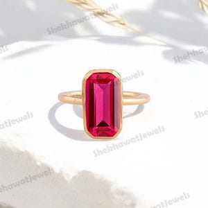 May include: A gold ring with a vibrant red rectangular gemstone. The ring is set on a white background.