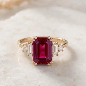 Puede incluir: Un anillo de oro con una gran piedra preciosa de color rojo intenso, de corte esmeralda rectangular en el centro. La piedra central está flanqueada a cada lado por una piedra rectangular transparente de corte baguette y una pequeña piedra redonda.