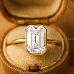 May include: A gold ring with a large, rectangular, clear gemstone set in a gold bezel. The ring is displayed in an open, brown, velvet-lined jewelry box. The ring's band is thin and gold.