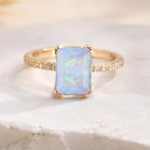 May include: A gold ring with a rectangular, light blue opal gemstone. The band is also gold and encrusted with small, clear gemstones. The ring is set on a white, textured surface.