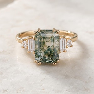 May include: A gold ring with a rectangular moss agate gemstone. The ring has a delicate band and is flanked by smaller, rectangular and round white gemstones. The moss agate has green and white patterns. The ring is set on a white surface.