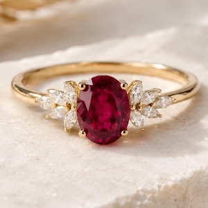 Op de afbeelding: Een gouden ring met een ovale robijn en marquise-geslepen diamanten. De band van de ring is van gepolijst goud. De robijn is dieprood en de diamanten schitteren. De ring ligt op een getextureerd, lichtgekleurd oppervlak.