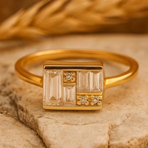 Könnte beinhalten: Ein goldener Ring mit rechteckiger Fassung, besetzt mit klaren, rechteckig geschliffenen Steinen und kleinen runden Akzentsteinen. Der Ring liegt auf einer hellen, strukturierten Oberfläche mit unscharfem Hintergrund.