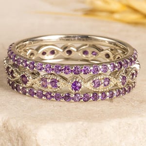 以下が含まれることがあります： 紫色の宝石が付いた3つのシルバーリング。 リングは積み重ねられており、複雑なデザインと鮮やかな色を示しています。 宝石は繰り返しのパターンでセットされており、きらめく効果を生み出しています。