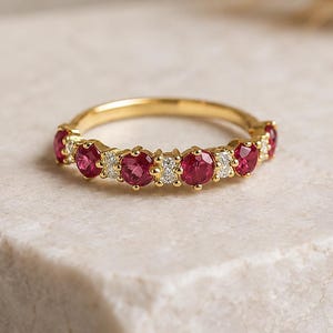 May include: A gold ring with alternating red and clear gemstones. The ring is set on a white and grey stone surface. The ring is a piece of jewellery.
