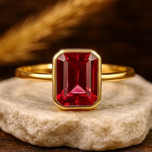 May include: A gold ring with a rectangular, deep red gemstone. The gemstone is set in a gold bezel, and the ring is displayed on a light-coloured, textured surface. The ring's band is a simple, polished gold.