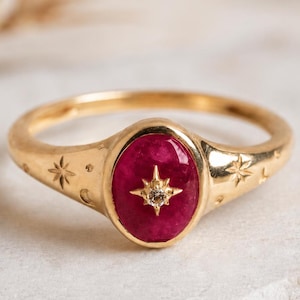 Pode incluir: Um anel de ouro com uma pedra oval de cor rosa profundo no centro. A pedra é acentuada com um pequeno detalhe central em forma de estrela. A banda de ouro é gravada com pequenos padrões de estrelas e pontos nas laterais do engaste.