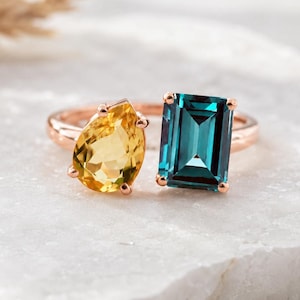 May include: A rose gold ring with two gemstones: a yellow, pear-shaped citrine and a rectangular teal gemstone. The ring sits on a white marble surface, highlighting the jewellery's design and colour contrast.