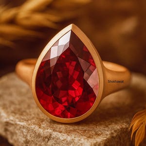 May include: A gold ring with a large, faceted, teardrop-shaped red gemstone. The ring is set on a textured, gray stone. The ring's design is simple, with a smooth gold band and bezel setting. The word "Shekhawat" is visible on the ring.