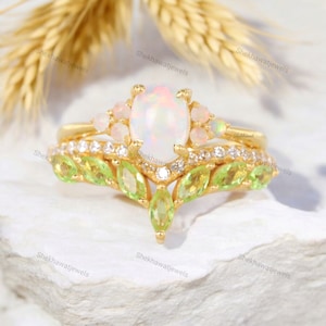 May include: A delicate gold ring set with a large oval opal, surrounded by smaller opals and sparkling clear stones. The band features a delicate design of green pear-shaped stones, resembling leaves.