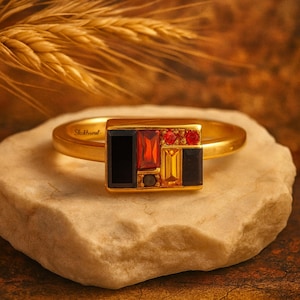 Può includere: Un anello d'oro con una faccia rettangolare con un mosaico di pietre preziose nere, rosse e gialle. L'anello è appoggiato su una pietra chiara, con uno sfondo sfocato di spighe di grano.