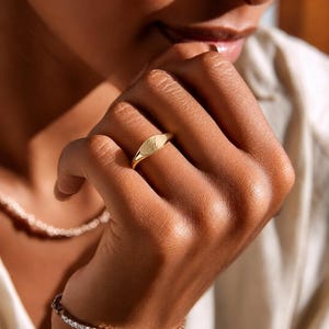 Op de afbeelding: Een gouden zegelring met de letter "A" gegraveerd op het ovale oppervlak. De ring wordt aan een vinger gedragen. De afbeelding is een close-up van de hand en de ring. Een diamanten armband en een ketting zijn ook zichtbaar.